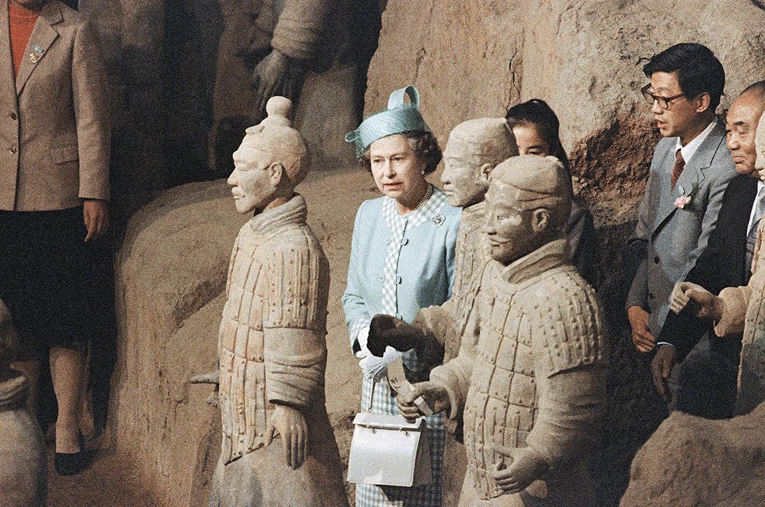 Queen Elizabeth stands between life-size terracotta soldiers, followed by Chinese dignitaries. The Queen wears a sky blue pillbox hat with a sheer veil and a matching sky blue suit jacket and dress with white and blue gingham edging.