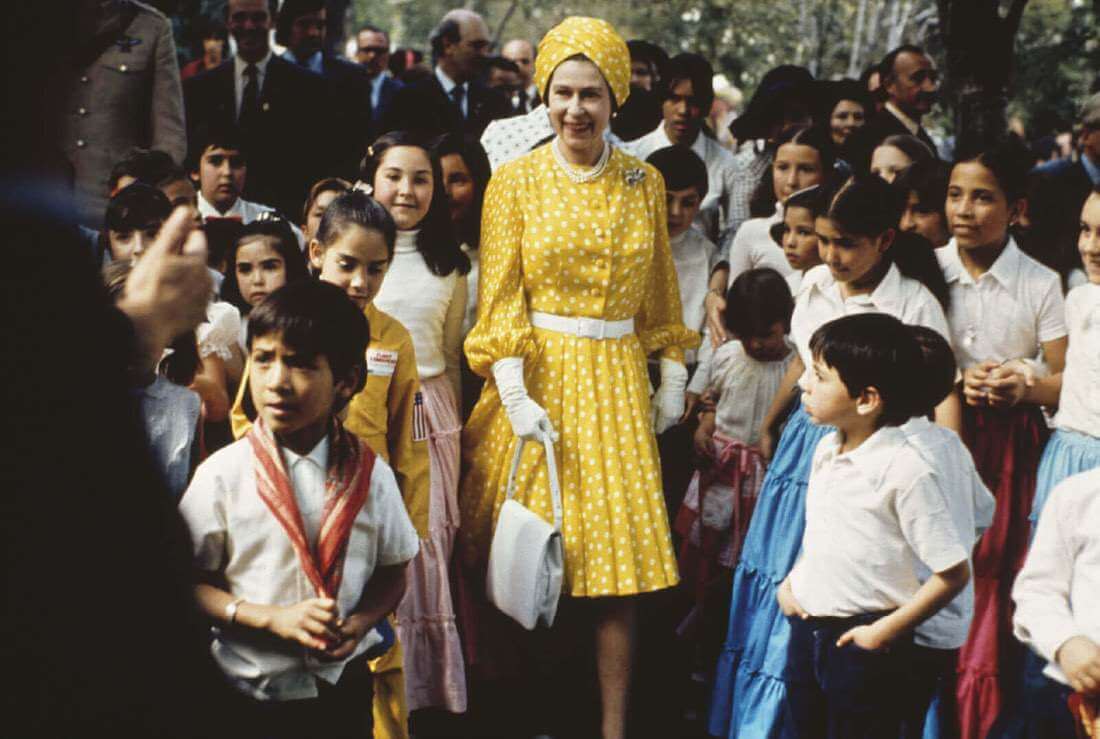 Queen Elizabeth stands out among a crowd of schoolchildren wearing a bright yellow turban-style hat and a matching polka dot dress fastened at the waist with a white belt.