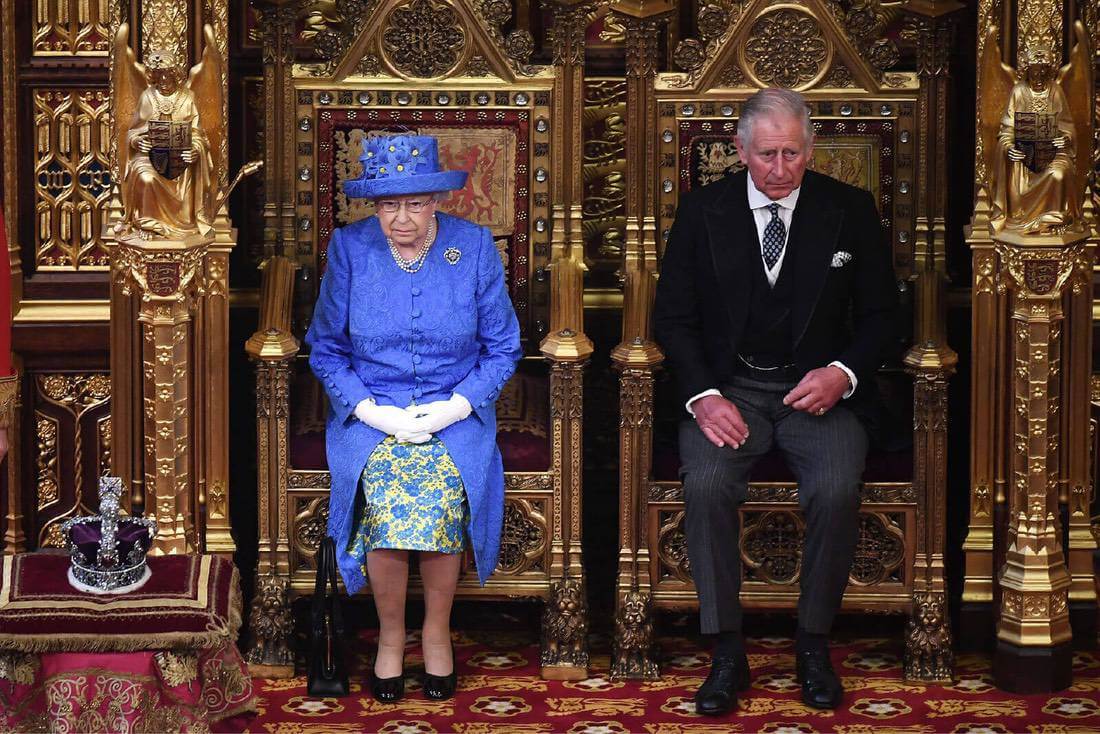 Queen Elizabeth sits on a golden throne in Parliament next to her son Prince Charles wearing a morning suit. The Queen wears a royal blue hat with yellow flower details and a matching button-up coat and dress.
