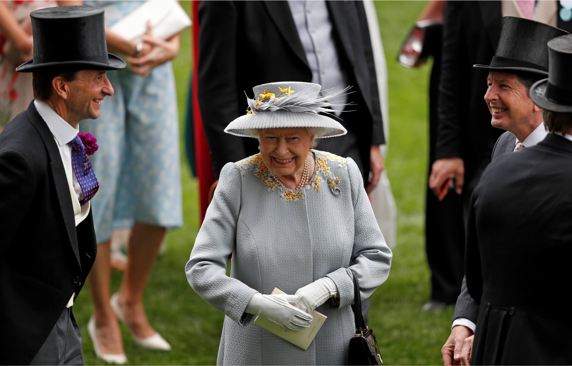 Queen Elizabeth wears a gray coat embroidered with yellow flowers around the collar, with a matching medium-brimmed gray hat decorated with feathers and yellow flowers. She is surrounded by men with black top hats and tails.
