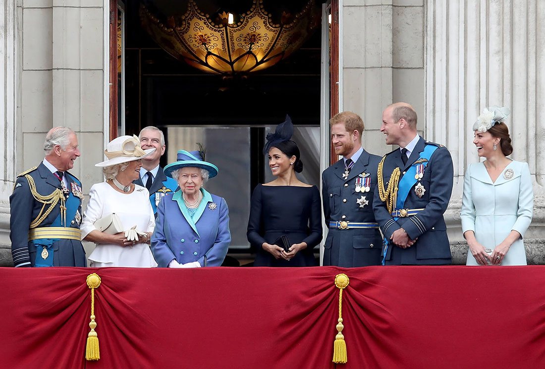 Queen Elizabeth stands on the balcony of Buckingham Palace, surrounded by senior members of the royal family, including Charles, Camilla, Meghan and Harry, all smiling at her. The Queen wears a royal blue and teal coat and matching hat with black feathers.