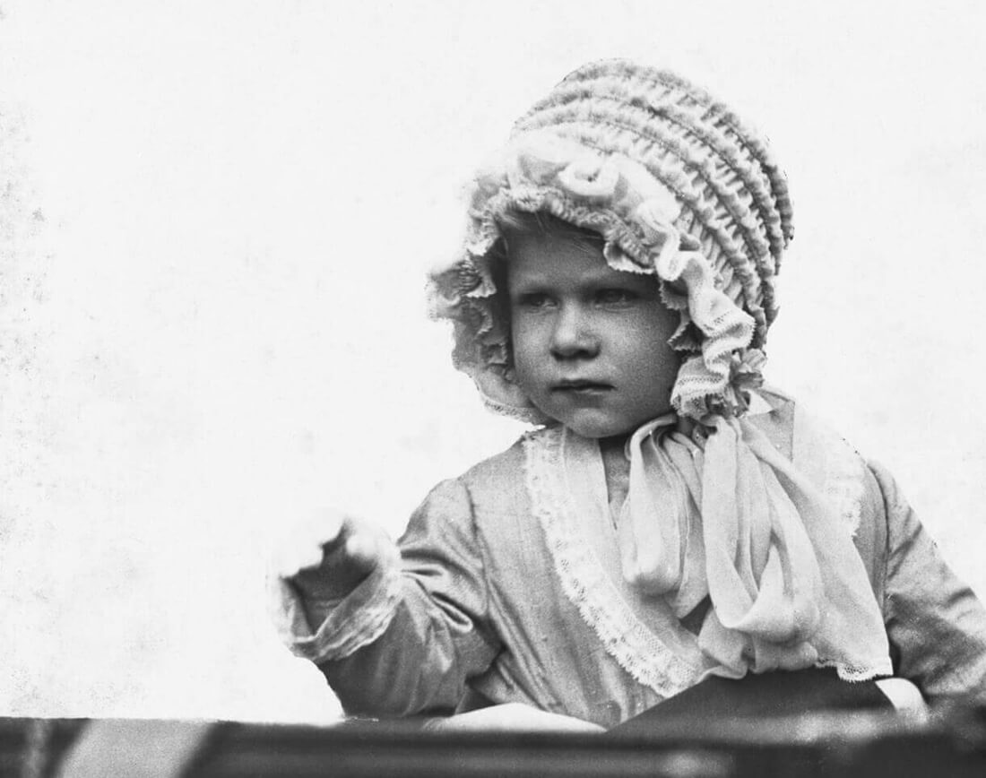 A baby Elizabeth sitting upright in a carriage wearing a detailed lace cap tied with a bow under her chin and a lace-trimmed cloak.