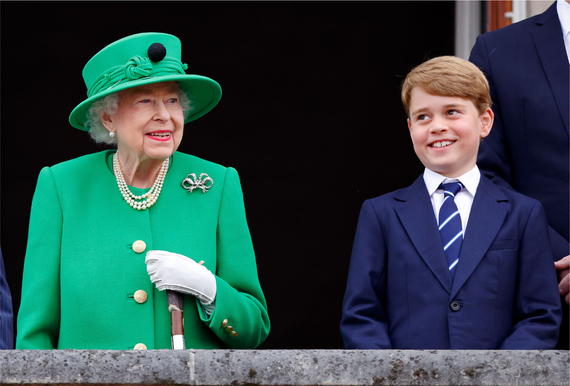 Queen Elizabeth stands on a balcony next to a smiling young Prince George. The Queen wears an emerald green suit jacket with a matching wide-brimmed hat, a three-string pearl necklace and a diamond brooch. She leans on a cane which she grips with a white gloved hand.