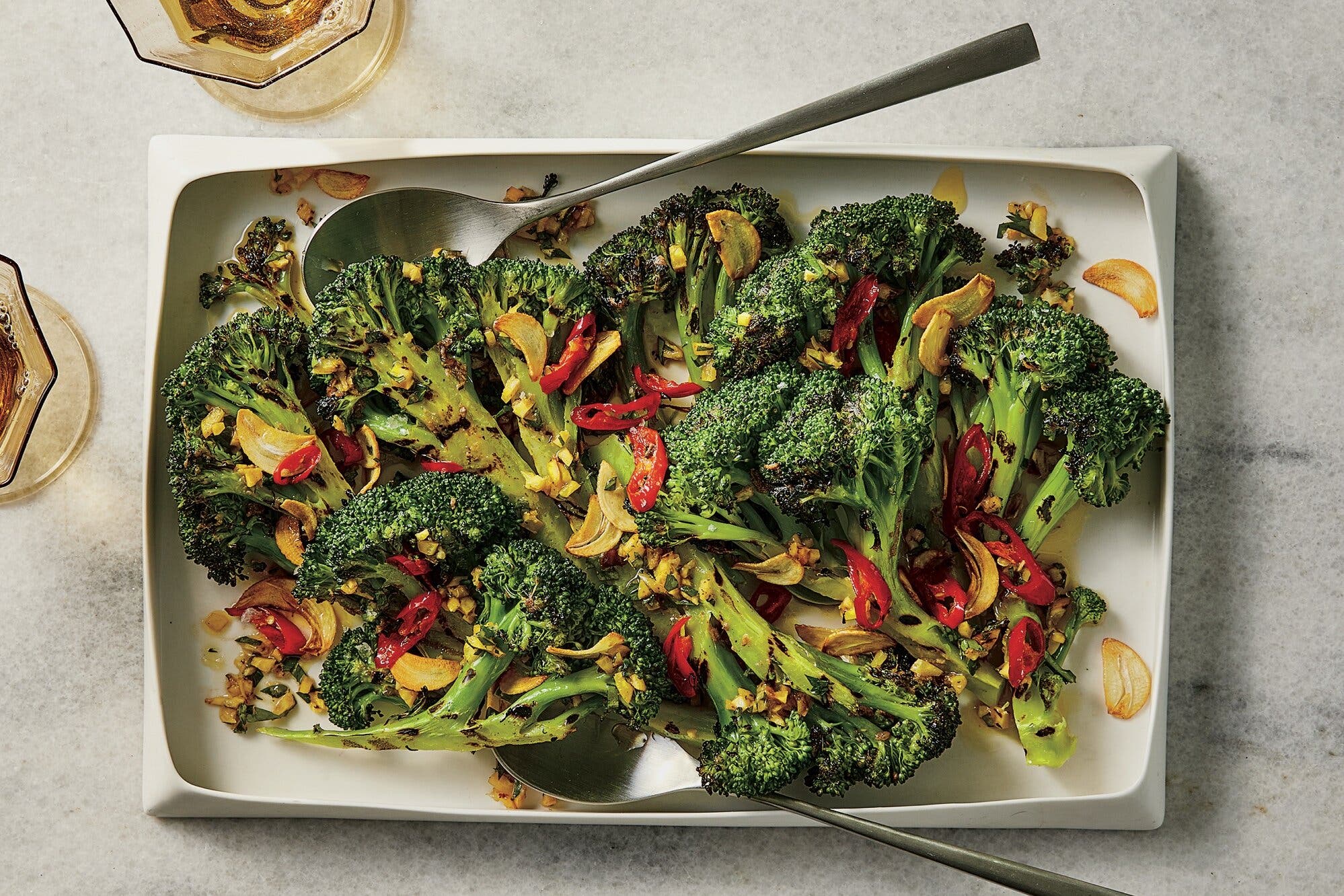 Broccoli garnished with sliced ​​garlic and pepper sits on a rectangular dish with a large metal spoon.